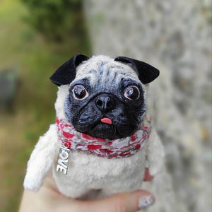 Puede incluir: Un peluche de perro carlino con hocico, orejas y ojos grandes negros. Lleva una bufanda estampada en rojo y blanco con un dije plateado con la palabra "LOVE". La lengua del carlino está afuera y tiene un cuerpo blanco.