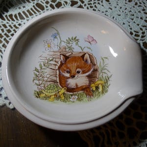 May include: A white ceramic spoon rest with a whimsical illustration of a fox peeking out of a log, surrounded by flowers and a turtle. The spoon rest has a curved shape with a small lip.