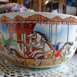 May include: White ceramic bowl with a carousel design. The bowl features a detailed illustration of carousel horses in various colors, including white, brown, and green, set against a backdrop of a carousel structure and a blue sky. The rim has a decorative gold pattern.