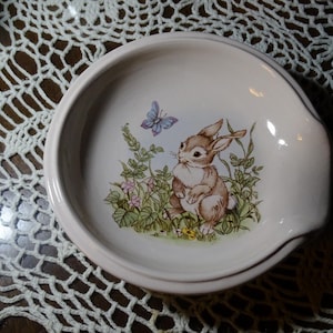 May include: A round, off-white ceramic spoon rest with a raised edge. The center features a charming illustration of a brown bunny, a blue butterfly, and wildflowers. The spoon rest has a small, curved handle.
