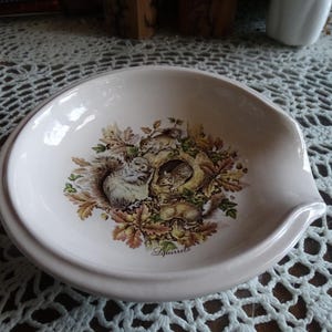 May include: A white ceramic spoon rest with a detailed illustration of squirrels nestled among autumn leaves. The word "Squirrels" is printed below the illustration. The spoon rest has a curved design.