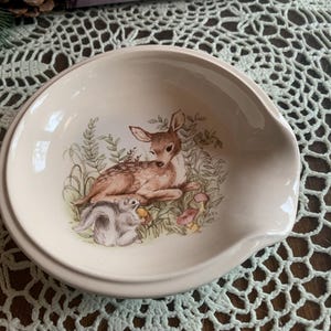 May include: A cream-colored ceramic spoon rest with a woodland scene featuring a fawn and squirrel. The spoon rest has a curved design and a raised edge. The illustration includes green foliage and mushrooms.