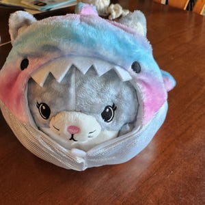 May include: A plush toy with a gray face and pink nose, wearing a blue, pink, and white shark costume.