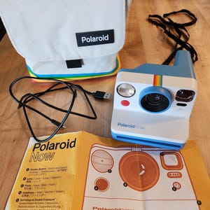 May include: A blue and white Polaroid Now instant camera with a black charging cable, a white carrying bag with a rainbow stripe, and an instructional pamphlet featuring a diagram of the camera labeled with the text Polaroid Now.