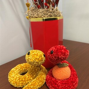 May include: Two crocheted snake plushies, one yellow and one red, with the red snake holding a crocheted orange. The snakes are sitting on a wooden surface.