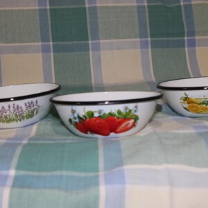 May include: Three white enamel bowls with black rims. The bowls feature different floral designs: lavender, strawberries, and lemons. The bowls are on a green and white checked tablecloth.