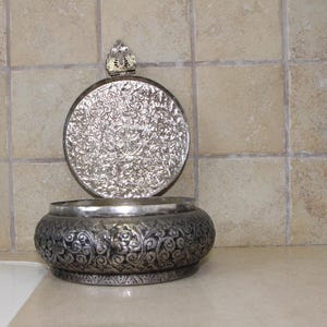 May include: A silver trinket box with an ornate floral design. The lid is open and has a small decorative knob on top.