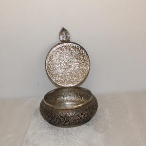 May include: A silver decorative bowl with an intricate floral design. The bowl has a lid with a small knob on top.