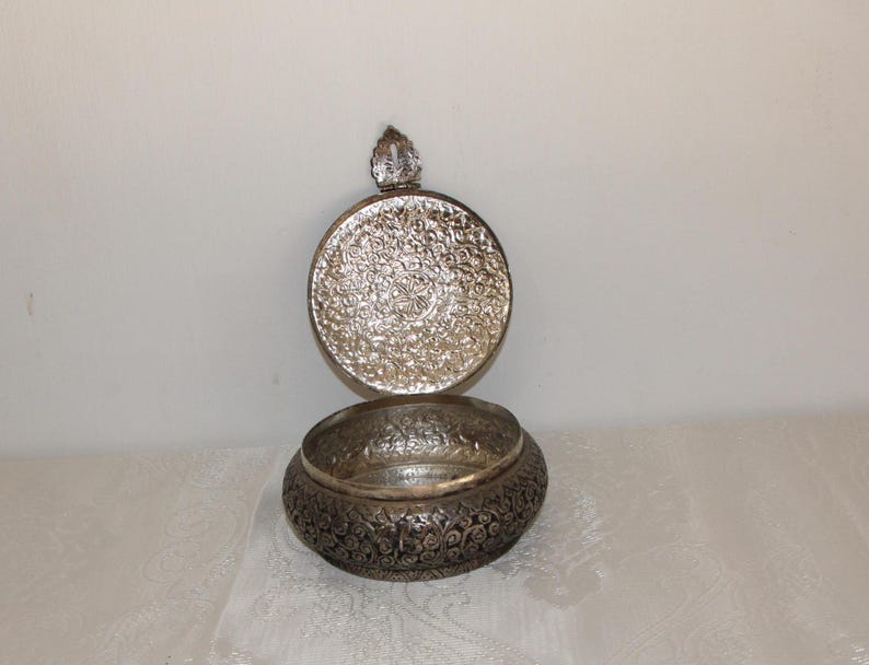 May include: A silver decorative bowl with an intricate floral design. The bowl has a lid with a small knob on top.