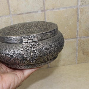 May include: A silver round trinket box with an ornate floral design and a hinged lid. The box has a decorative clasp.
