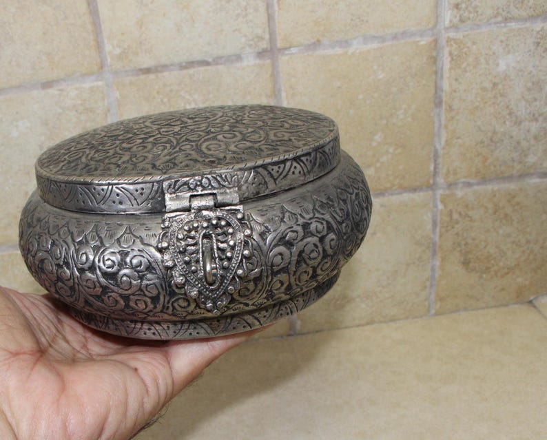 May include: A silver round trinket box with an ornate floral design and a hinged lid. The box has a decorative clasp.