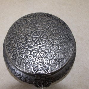May include: A round silver metal box with an intricate floral design embossed on the lid. The box has a hinged lid and a small metal handle.