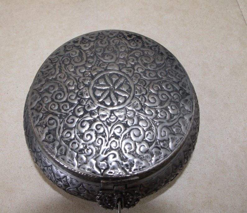 May include: A round silver metal box with an intricate floral design embossed on the lid. The box has a hinged lid and a small metal handle.