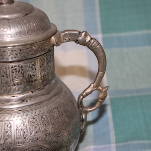 Puede incluir: Una jarra de plata con intrincados diseños grabados y una asa decorativa. La jarra tiene una tapa y está hecha de metal.
