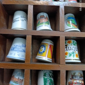 Puede incluir: Una colección de dedales decorativos exhibidos en un organizador de madera. Los dedales son blancos con varios diseños coloridos, incluyendo paisajes y edificios. El organizador tiene múltiples compartimentos, cada uno con un dedal.