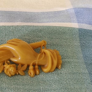 May include: A vintage brooch made of brown plastic, depicting a hand holding a bouquet of flowers. The flowers are stylised and have a simple design.