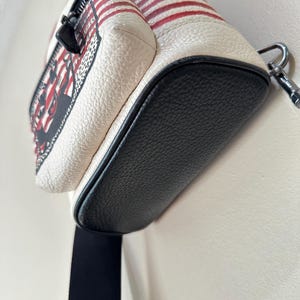 Puede incluir: Un bolso bandolera peque&ntilde;o con cuerpo de cuero texturizado en color blanco roto, con rayas horizontales rojas y una base de cuero negro. El bolso incluye una cremallera negra, un detalle superior de cuero rojo y una correa de tela negra con un clip de metal plateado.