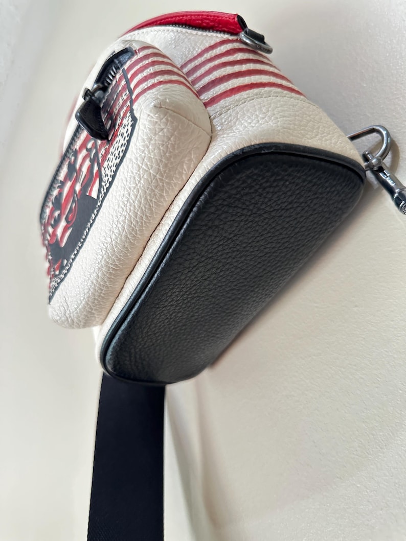 Puede incluir: Un bolso bandolera peque&ntilde;o con cuerpo de cuero texturizado en color blanco roto, con rayas horizontales rojas y una base de cuero negro. El bolso incluye una cremallera negra, un detalle superior de cuero rojo y una correa de tela negra con un clip de metal plateado.