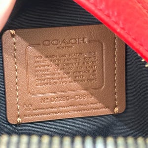 Puede incluir: Un parche rectangular de cuero color canela dentro de un bolso rojo con forro negro. El parche presenta el texto en relieve COACH NEW YORK, seguido de una descripci&oacute;n de la colaboraci&oacute;n de Keith Haring y Disney, el n&uacute;mero D2280-C6910 y texto de copyright.