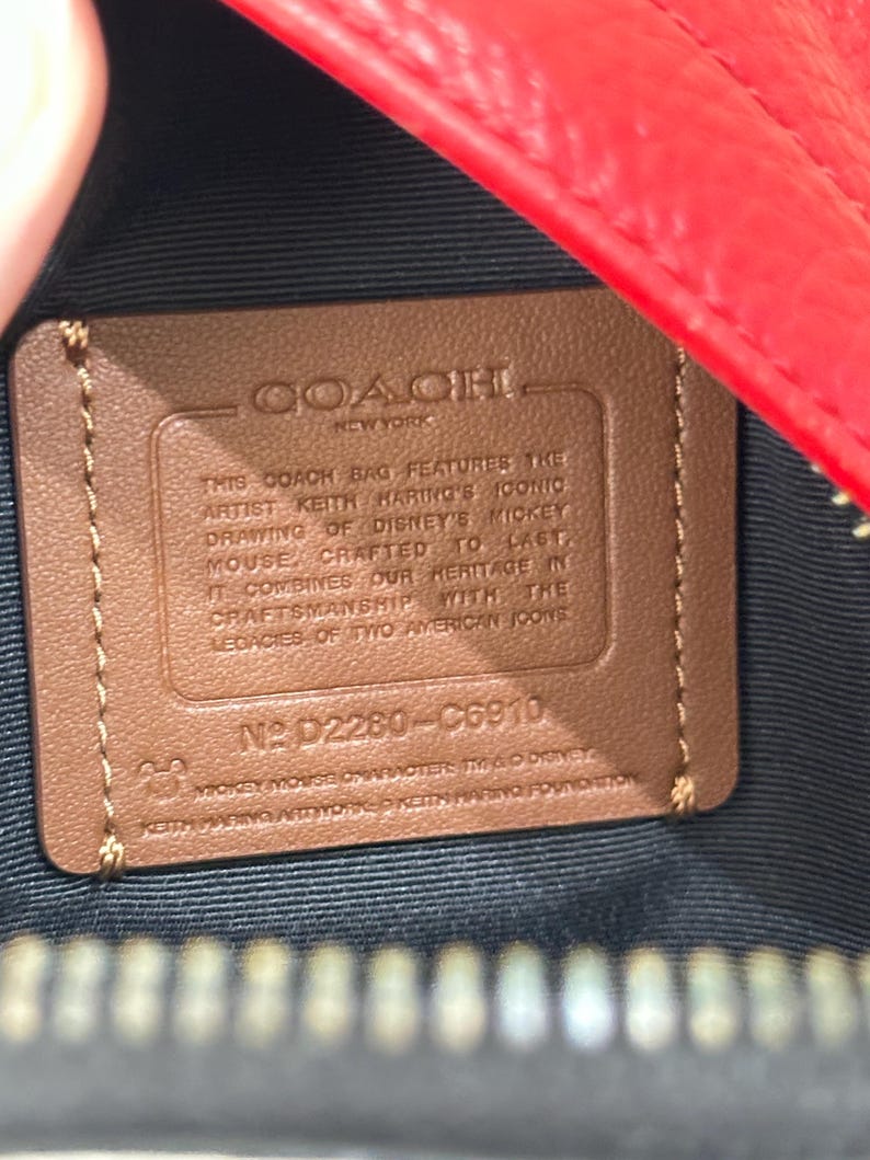 Puede incluir: Un parche rectangular de cuero color canela dentro de un bolso rojo con forro negro. El parche presenta el texto en relieve COACH NEW YORK, seguido de una descripci&oacute;n de la colaboraci&oacute;n de Keith Haring y Disney, el n&uacute;mero D2280-C6910 y texto de copyright.