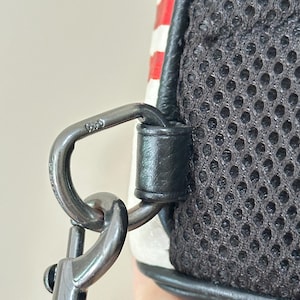 Puede incluir: Un accesorio de anillo en D de metal negro en un bolso, asegurado por una presilla de cuero negro. El bolso presenta un panel trasero de malla negra y una secci&oacute;n a rayas rojas y blancas. Un mosquet&oacute;n giratorio de metal negro est&aacute; enganchado.