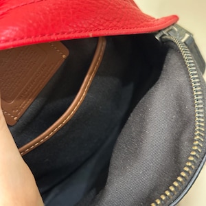 Puede incluir: El interior de un bolso de cuero rojo con forro de tela negra, cierre de cremallera met&aacute;lica y un parche de cuero marr&oacute;n con texto grabado. El bolso tiene una superficie exterior texturizada y un borde de cuero marr&oacute;n en el bolsillo interior.