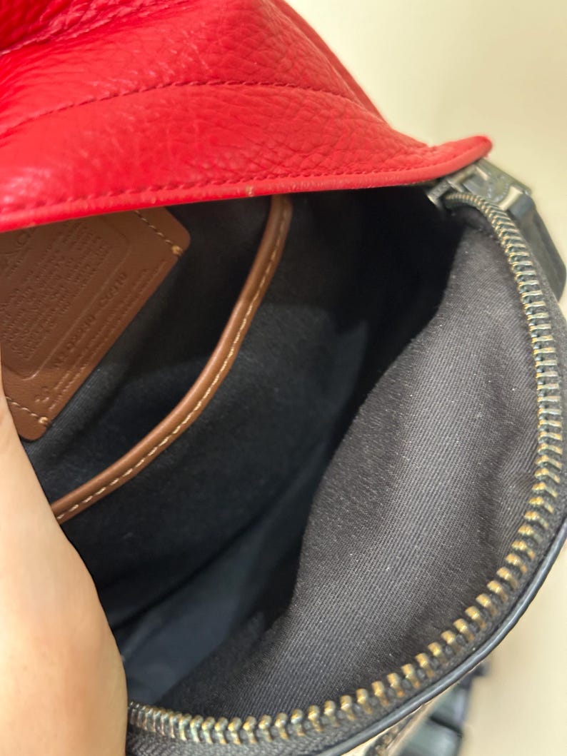 Puede incluir: El interior de un bolso de cuero rojo con forro de tela negra, cierre de cremallera met&aacute;lica y un parche de cuero marr&oacute;n con texto grabado. El bolso tiene una superficie exterior texturizada y un borde de cuero marr&oacute;n en el bolsillo interior.