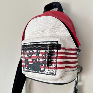Puede incluir: Una mochila peque&ntilde;a de cuero texturizado en rojo y blanco con un bolsillo frontal con cremallera que presenta una ilustraci&oacute;n gr&aacute;fica en blanco y negro de un personaje de dibujos animados. La bolsa tiene rayas rojas horizontales.