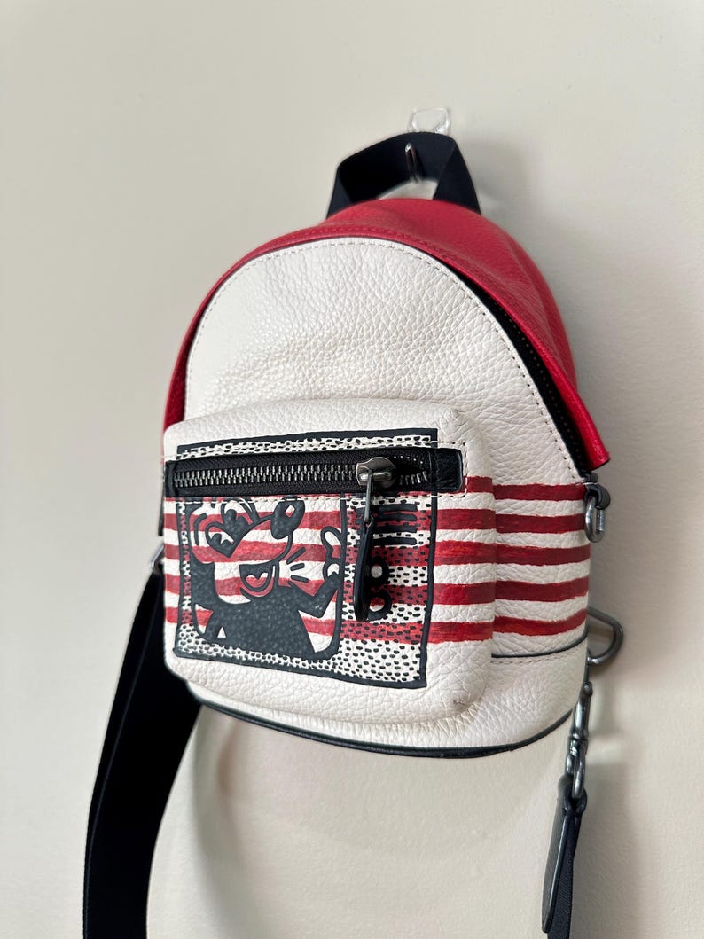 Puede incluir: Una mochila peque&ntilde;a de cuero texturizado en rojo y blanco con un bolsillo frontal con cremallera que presenta una ilustraci&oacute;n gr&aacute;fica en blanco y negro de un personaje de dibujos animados. La bolsa tiene rayas rojas horizontales.