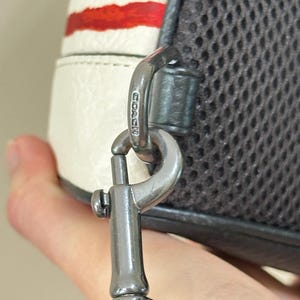 Puede incluir: Un mosquet&oacute;n de metal gris oscuro unido a un bolso blanco con rayas horizontales rojas. La pieza de metal tiene grabado el nombre COACH. El bolso cuenta con un panel de malla gris oscuro en el lateral, conectado al mosquet&oacute;n por una trabilla de cuero.