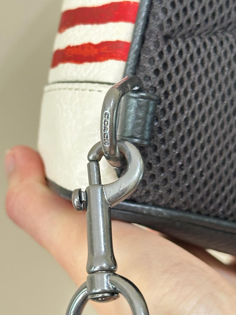 Puede incluir: Un mosquet&oacute;n de metal gris oscuro unido a un bolso blanco con rayas horizontales rojas. La pieza de metal tiene grabado el nombre COACH. El bolso cuenta con un panel de malla gris oscuro en el lateral, conectado al mosquet&oacute;n por una trabilla de cuero.