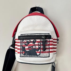 Puede incluir: Una mochila peque&ntilde;a de color blanco con rayas horizontales rojas y una solapa superior roja. El bolsillo frontal presenta un gr&aacute;fico en blanco y negro de un personaje de dibujos animados con nariz y orejas grandes, con cremallera negra.