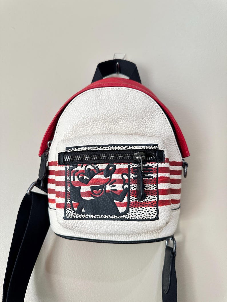 Puede incluir: Una mochila peque&ntilde;a de color blanco con rayas horizontales rojas y una solapa superior roja. El bolsillo frontal presenta un gr&aacute;fico en blanco y negro de un personaje de dibujos animados con nariz y orejas grandes, con cremallera negra.