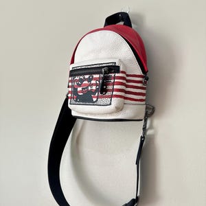 Puede incluir: Una mochila peque&ntilde;a con la parte superior roja y el cuerpo blanco de textura granulada con rayas rojas horizontales. El bolsillo frontal muestra un gr&aacute;fico en blanco y negro de un personaje de dibujos animados. Incluye una correa negra ajustable.