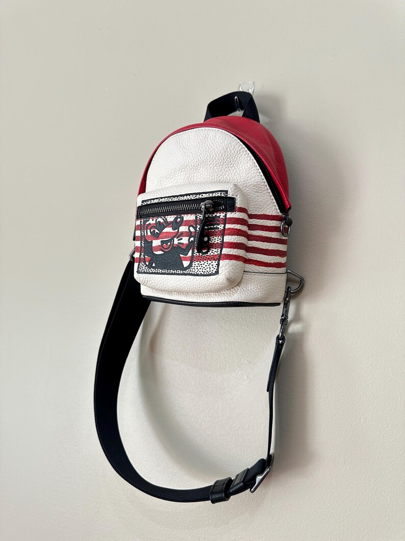 Puede incluir: Una mochila peque&ntilde;a con la parte superior roja y el cuerpo blanco de textura granulada con rayas rojas horizontales. El bolsillo frontal muestra un gr&aacute;fico en blanco y negro de un personaje de dibujos animados. Incluye una correa negra ajustable.