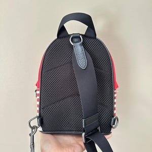 Puede incluir: Una mochila peque&ntilde;a con panel trasero de malla negra, asa superior y una sola correa de hombro negra ajustable. Los laterales de la bolsa muestran un patr&oacute;n de rayas rojas y blancas, e incluye anillas de sujeci&oacute;n de metal.