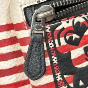Puede incluir: Un bolso de cuero texturizado blanco con rayas rojas horizontales y un dise&ntilde;o gr&aacute;fico negro. Un tirador de cremallera de cuero negro est&aacute; unido a un deslizador de metal plateado con el texto grabado Coach New York visible en el herraje.