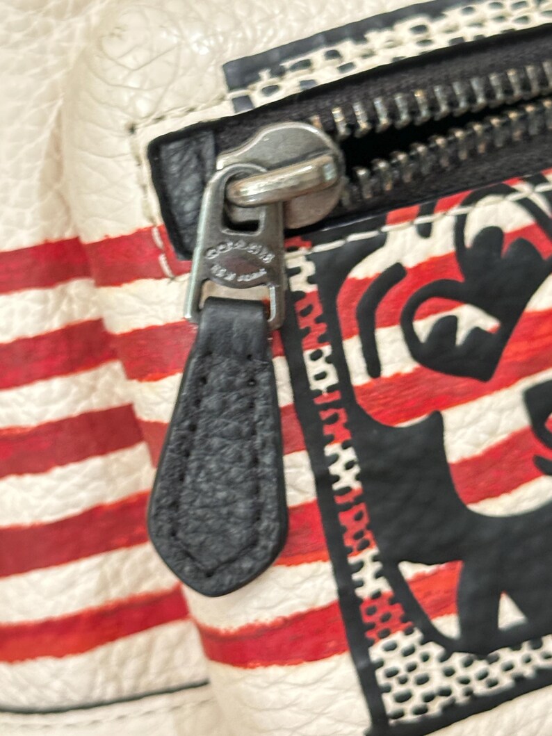 Puede incluir: Un bolso de cuero texturizado blanco con rayas rojas horizontales y un dise&ntilde;o gr&aacute;fico negro. Un tirador de cremallera de cuero negro est&aacute; unido a un deslizador de metal plateado con el texto grabado Coach New York visible en el herraje.