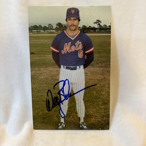 May include: A baseball card featuring a baseball player wearing a blue and orange Mets jersey with the number 6 on the front. The player is standing on a baseball field and has a blue signature on the card.