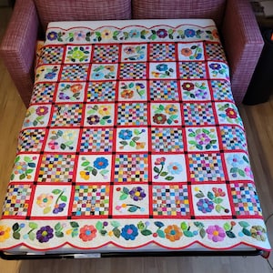 May include: A white quilt with a red border and a scalloped edge. The quilt is made up of squares with a variety of floral patterns in shades of pink, blue, green, and yellow. The quilt is displayed on a couch.