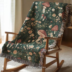 May include: A dark green throw blanket featuring a botanical design of mushrooms and flowers, draped over a wooden rocking chair. The blanket has a fringed edge and the text "TheCozyTales" is visible.