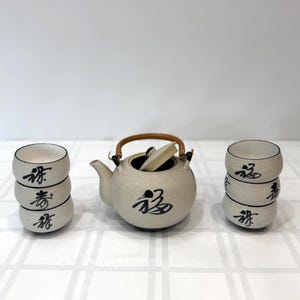 Puede incluir: Juego de té de cerámica beige con caligrafía negra. El juego incluye una tetera con asa tejida y tapa, y seis tazas de té pequeñas. Las tazas y la tetera tienen caligrafía negra. El juego está sobre una superficie blanca.
