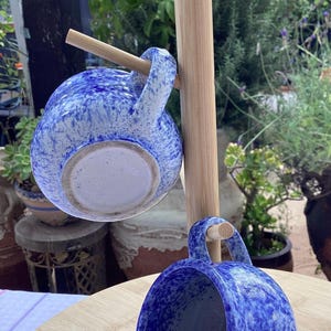 Puede incluir: Dos tazas de cerámica azul y blanca moteada colgadas de un árbol de tazas de madera. Las tazas tienen forma redondeada con asa. El fondo muestra un jardín con plantas y una mesa de madera.