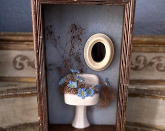 May include: A framed miniature scene of a white porcelain pedestal sink with blue flowers, a small oval mirror, and dried floral accents on a blue background.