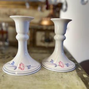 Puede incluir: Un par de candelabros de cerámica blanca con un diseño floral. Los candelabros tienen una base acampanada y un tallo delgado. El diseño floral presenta flores rosas, azules y moradas.