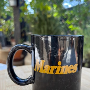 May include: Black coffee mug with the word "Marines" in yellow lettering. The mug has a large handle and sits on a wooden surface. The background is blurred with green foliage.