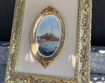 May include: An ornate gold-colored framed painting with an oval-shaped landscape scene. The painting depicts a coastal town with a mountain in the background. The frame has intricate detailing and is displayed on a wooden easel.