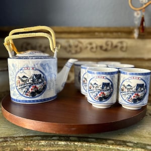 Op de afbeelding: Een blauwe en witte porseleinen theepot met een bamboe handvat en bijpassende theekopjes. De theepot en kopjes hebben een schilderachtig ontwerp van een bergdorp met een brug en bomen.