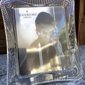Puede incluir: Un marco de fotos de cristal transparente con intrincados patrones tallados a lo largo del borde y esquinas redondeadas. El marco presenta el logotipo de Waterford Crystal en la parte superior central y contiene una foto en blanco y negro.