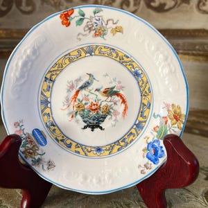Puede incluir: Un plato de porcelana blanca con borde azul y diseños intrincados de flores y pájaros. El plato presenta una imagen central de dos pájaros posados cerca de un arreglo floral en un jarrón decorativo. El plato está sostenido por un soporte de madera rojo.